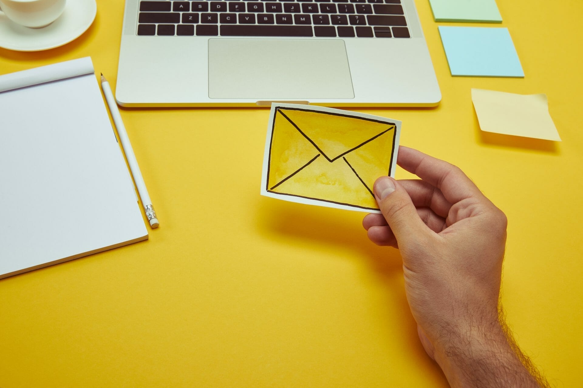 A hand holding a yellow envelope illustration next to a laptop, notebook, pencil, and sticky notes on a yellow surface.