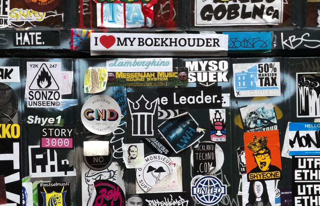 A wall covered with various colorful stickers and posters, including a prominent sticker reading "I ❤️ MY BOEKHOUDER.