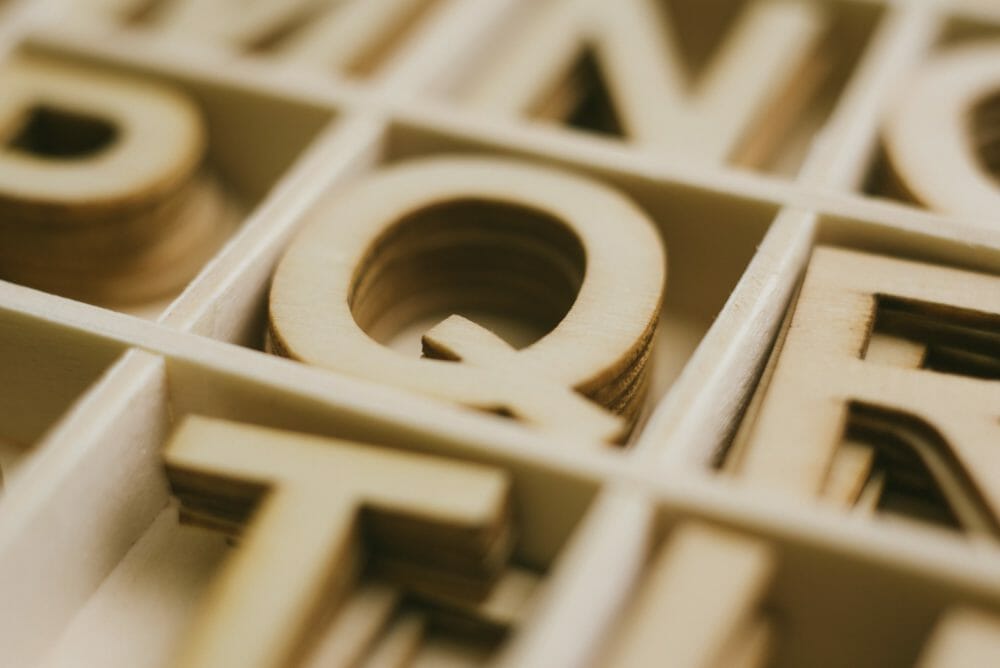 Close-up view of wooden letters stacked in a box, focusing on the letter "Q" in the center.