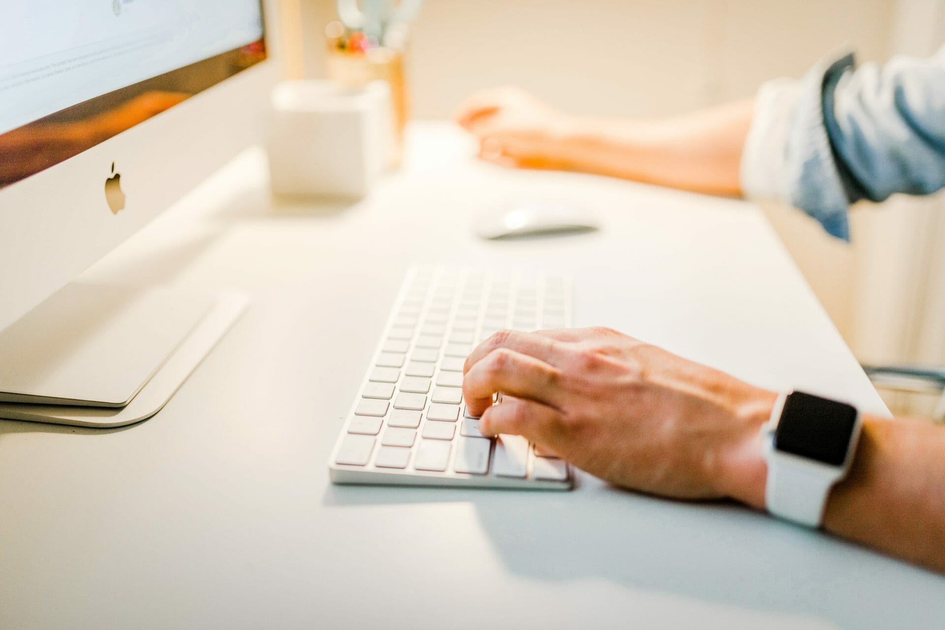 A person uses a desktop computer, typing on a wireless keyboard, and wearing a smartwatch.