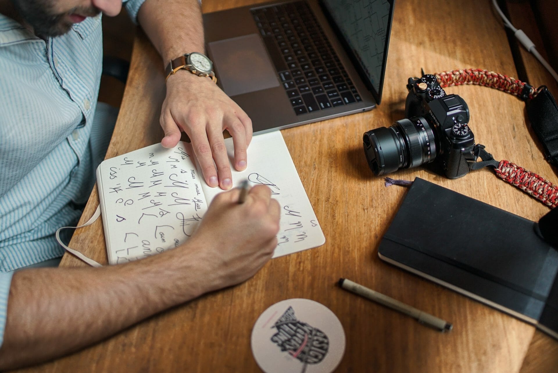 Person writing in a notebook with an open laptop, camera, and other items on a wooden desk.