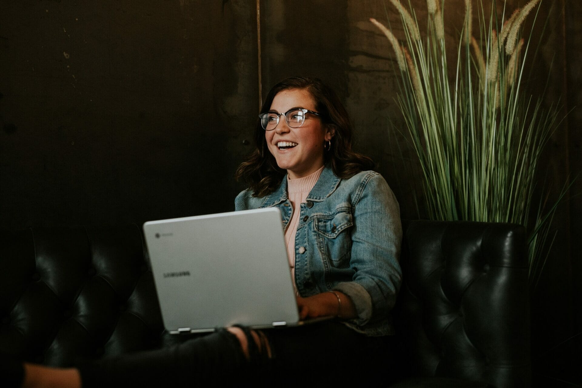 A person with glasses and a denim jacket is sitting on a leather couch, smiling while using a laptop.