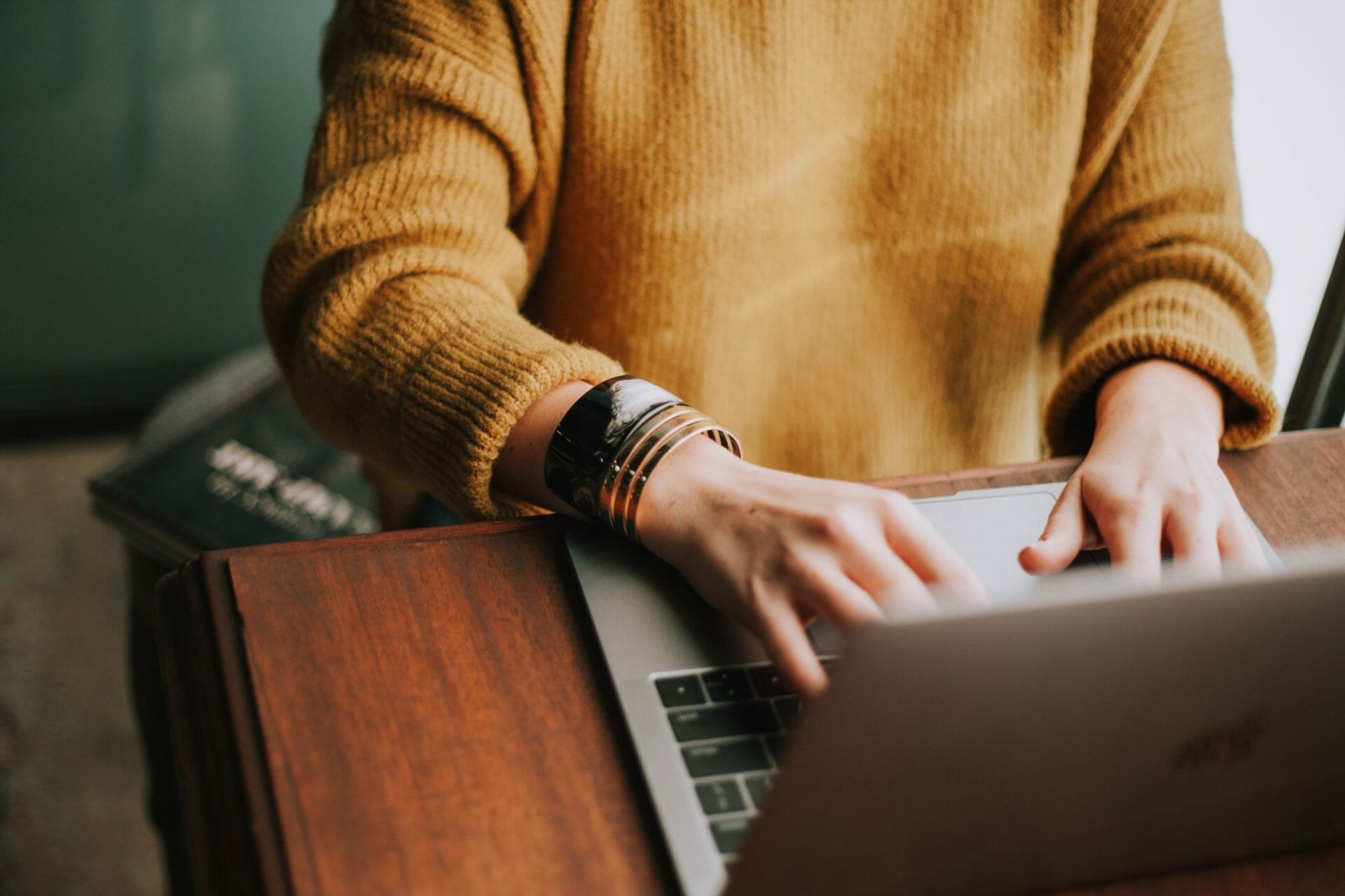 Person in a yellow sweater typing on a laptop, with a book partially visible on the table nearby.