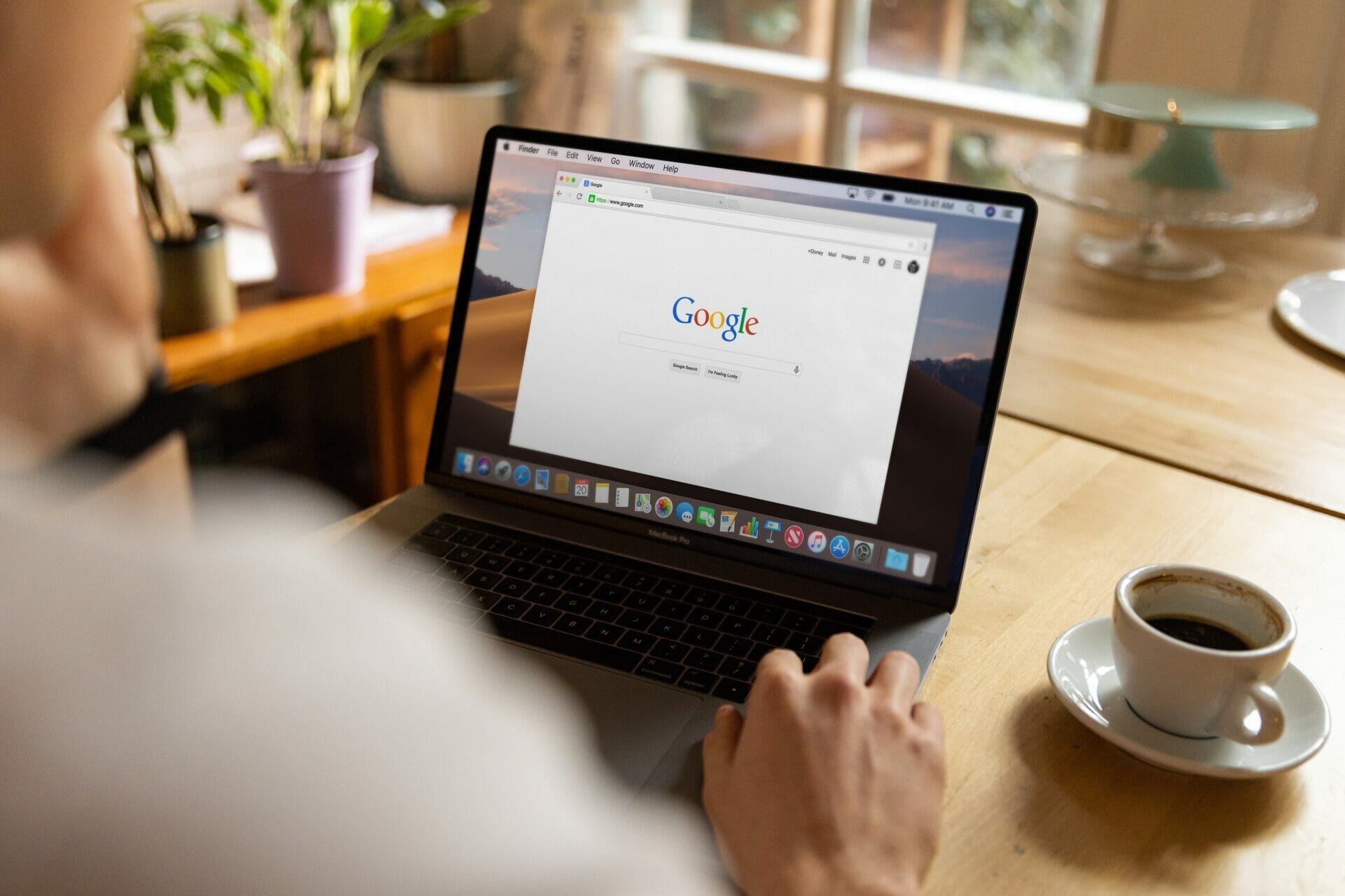 A person using a laptop with Google's search page open, sitting at a wooden table with a cup of coffee and potted plants in the background.