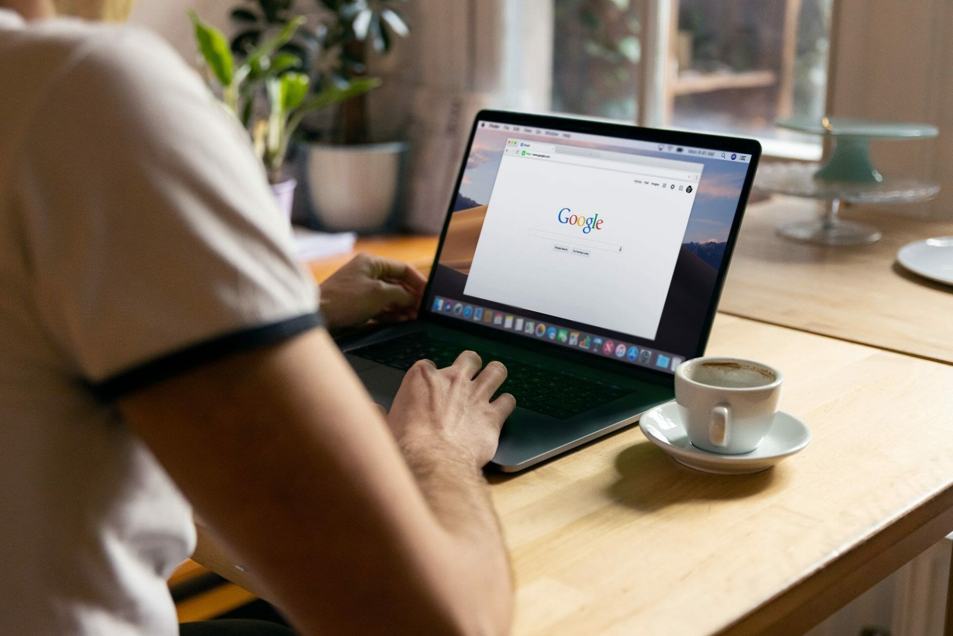 Person using a laptop with Google search engine open, sitting at a wooden table with a cup of coffee nearby and plants in the background.