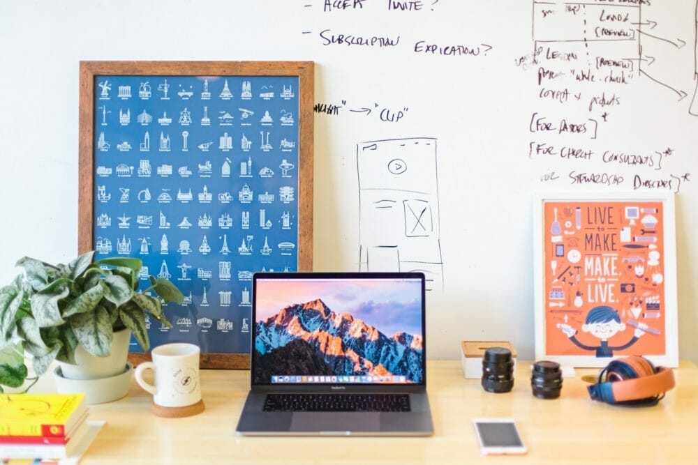 A cluttered desk with a laptop displaying a mountain scene, plant, books, headphones, camera lenses, coffee mug, and various posters. The whiteboard in the background has handwritten notes and diagrams.