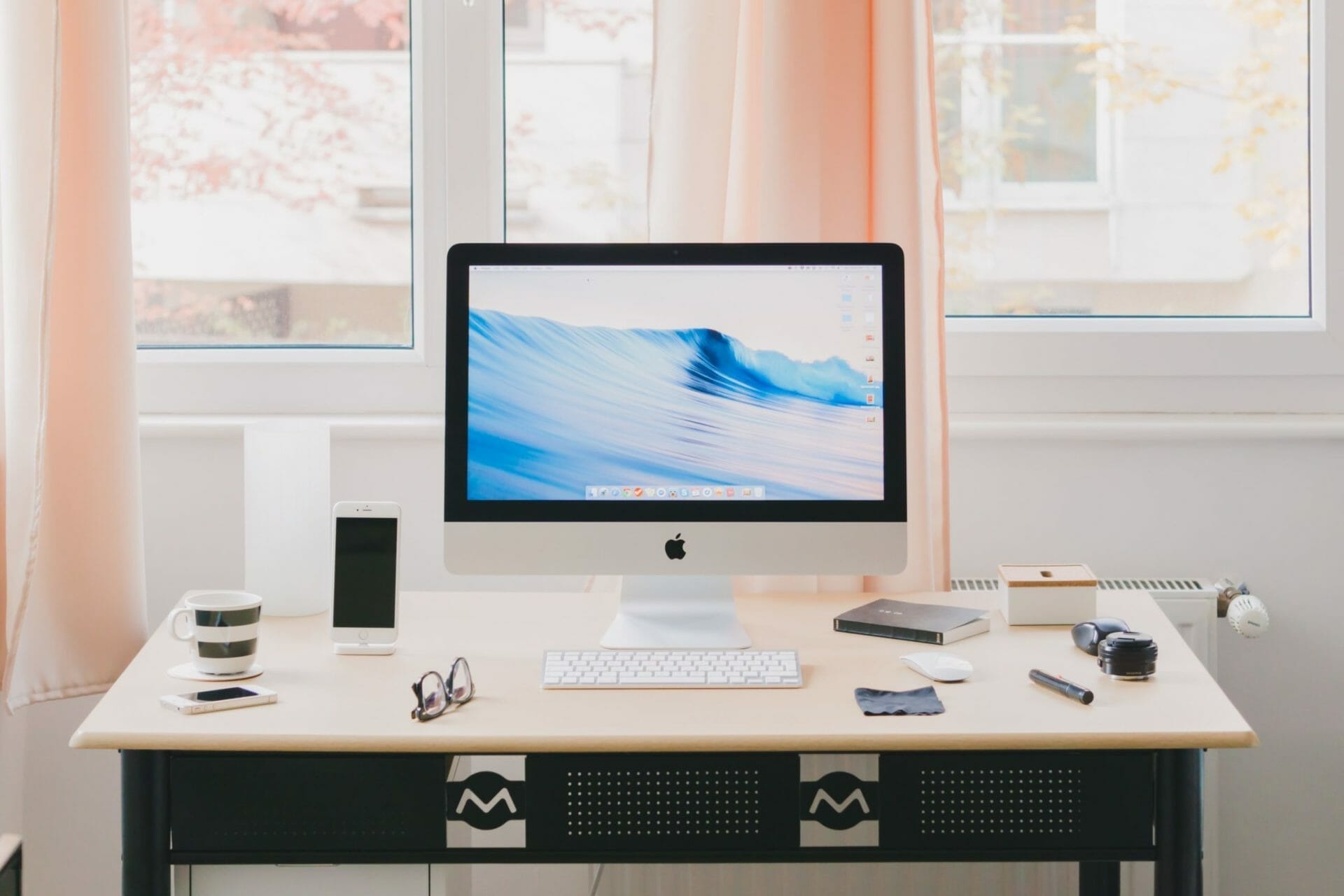 A tidy desk with an iMac displaying a wave wallpaper, a smartphone, various small items, and a window with peach curtains in the background.