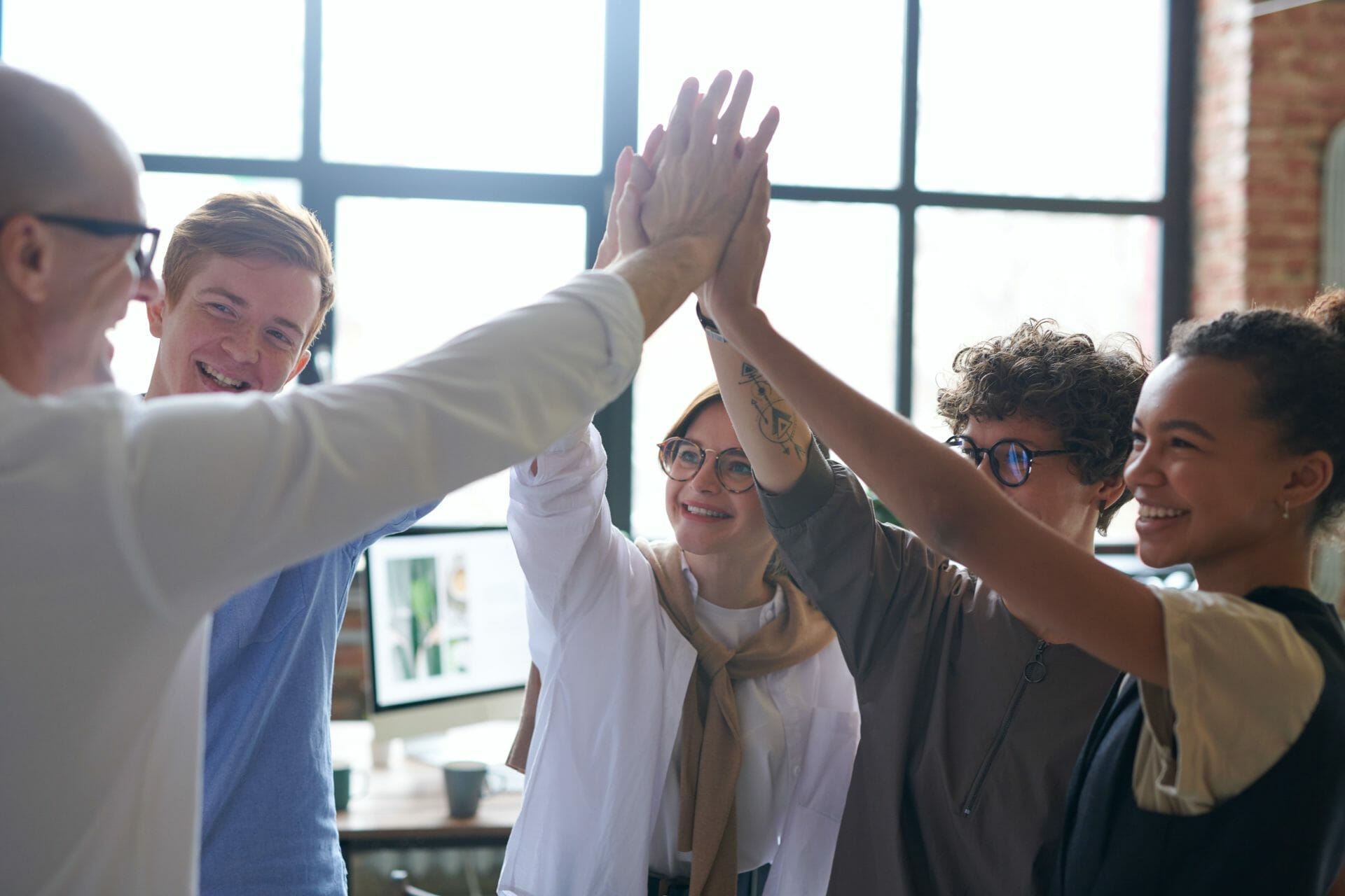 A group of five people, standing in a circle, joyfully raise their hands together in a high-five, inside a well-lit room with large windows.