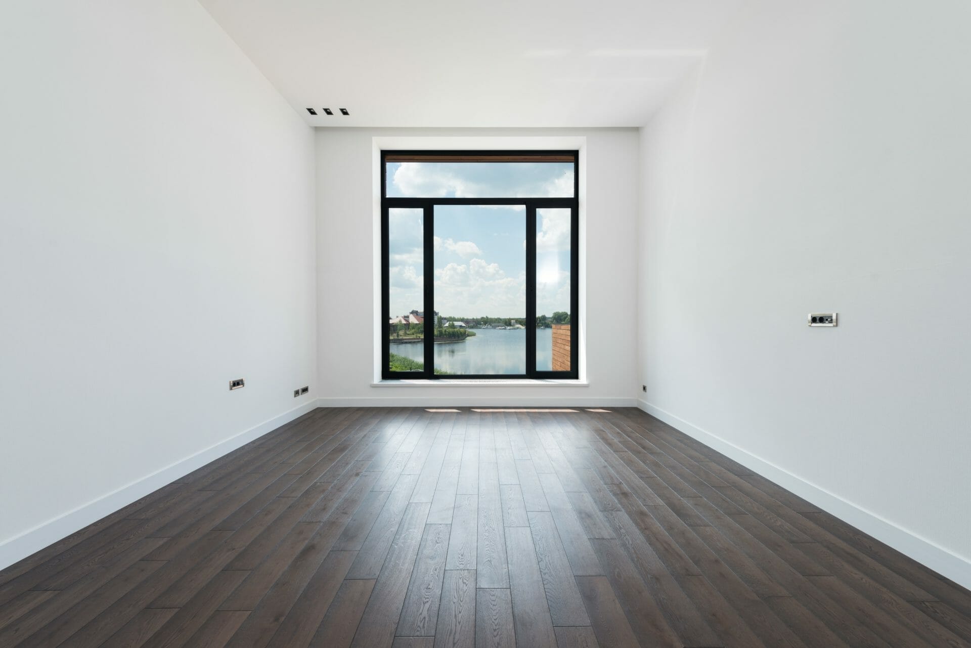 An empty room with a large window overlooking a lake, featuring dark wood flooring and white walls.