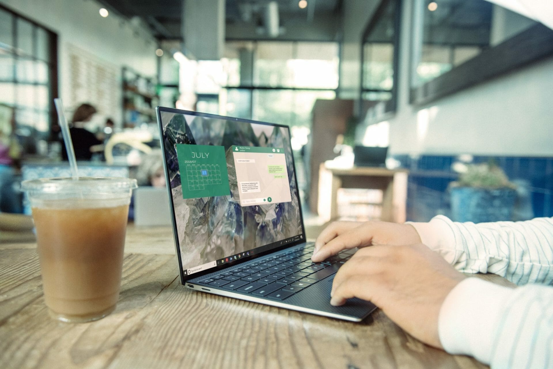 Person typing on a laptop at a wooden table with a plastic cup of iced coffee, in a modern café setting.