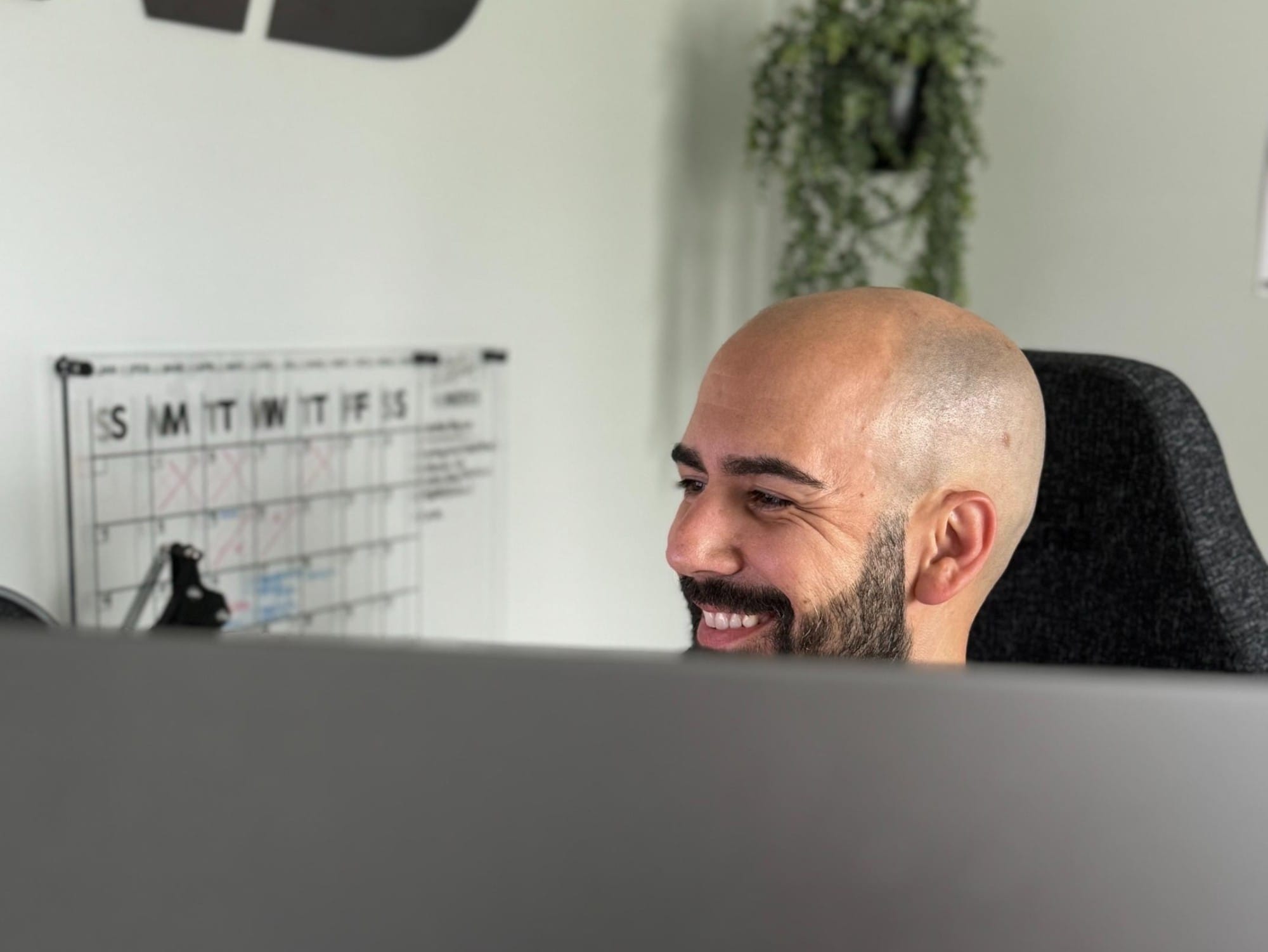 A man with a bald head and beard smiles while working at a desk, with a weekly calendar and plant visible in the background.