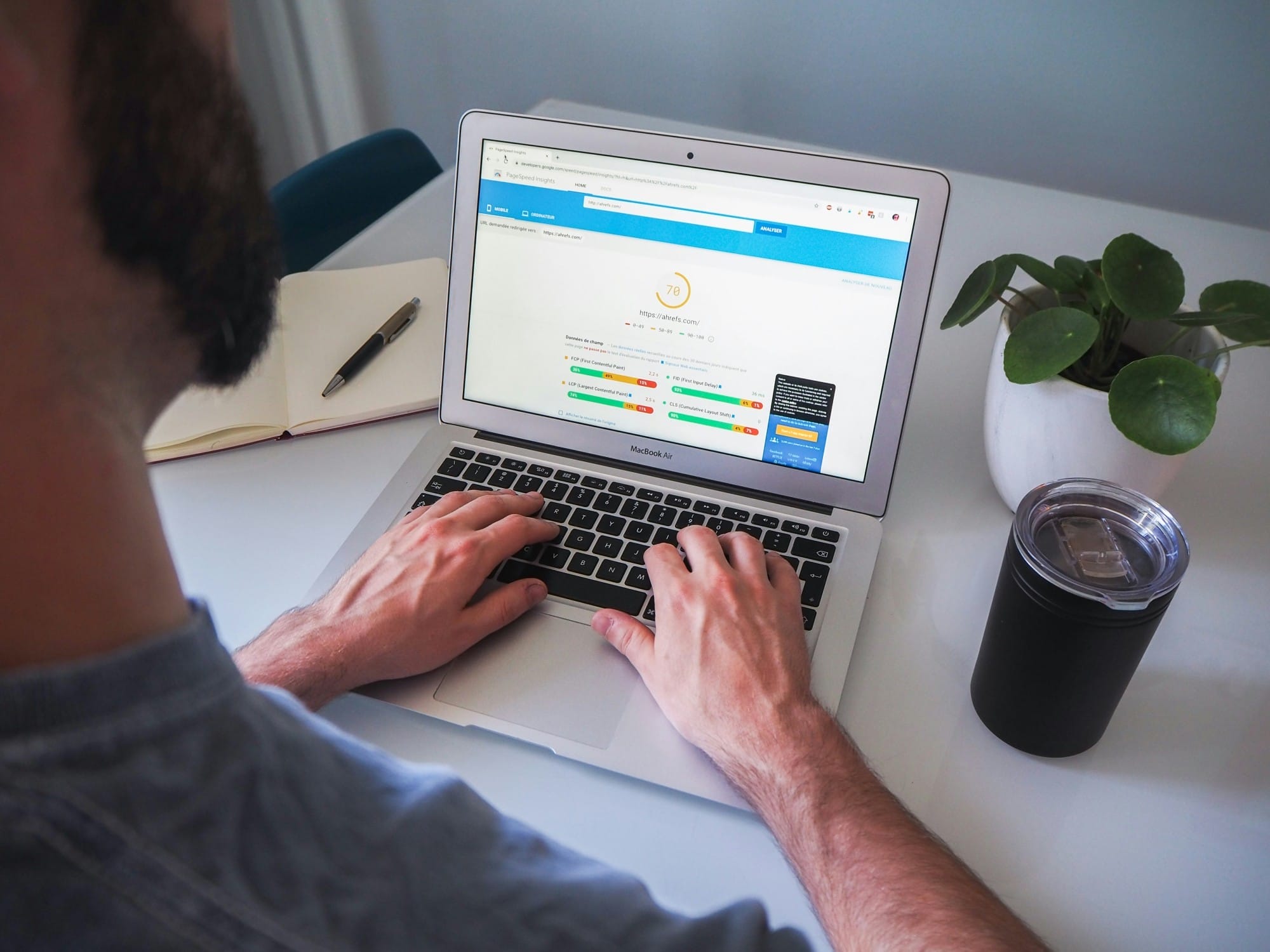 Person typing on a laptop displaying a webpage, with a notebook, pen, plant, and tumbler on a white desk.