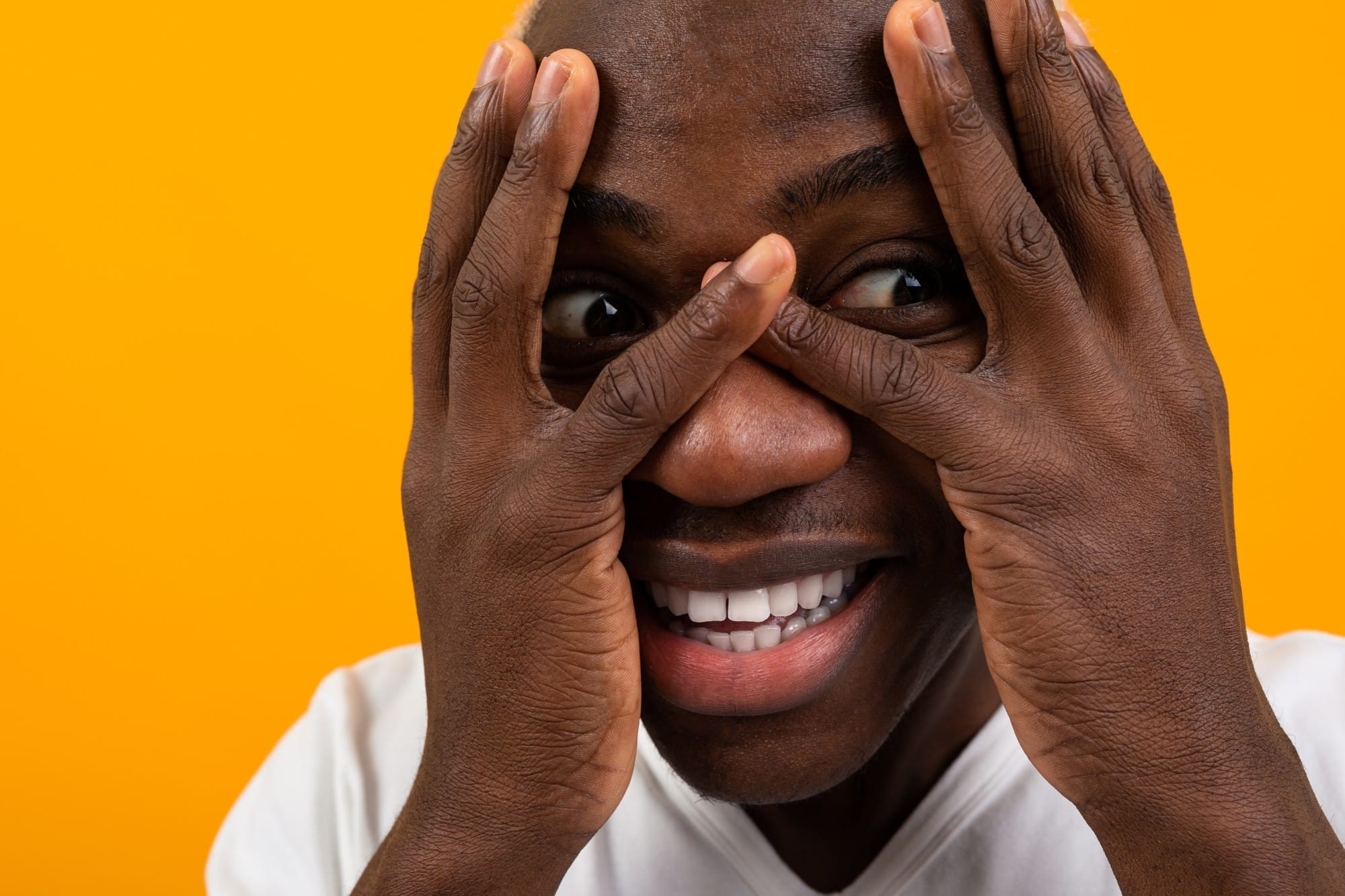 Person smiling while playfully peeking through fingers, against an orange background.
